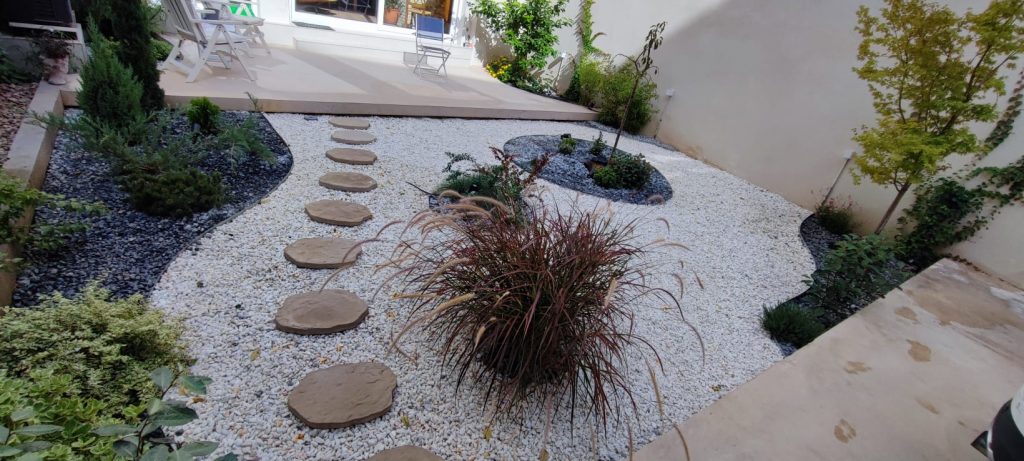 Jardín diseñado con grava blanca, sendero de piedras y áreas verdes con arbustos y plantas decorativas